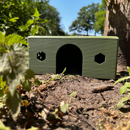 HexaHouse van olive groen kleurig PLA, geplaatst in een natuurlijke buitenomgeving – 3D-geprint knaagdierhuisje met zeshoekige ramen.