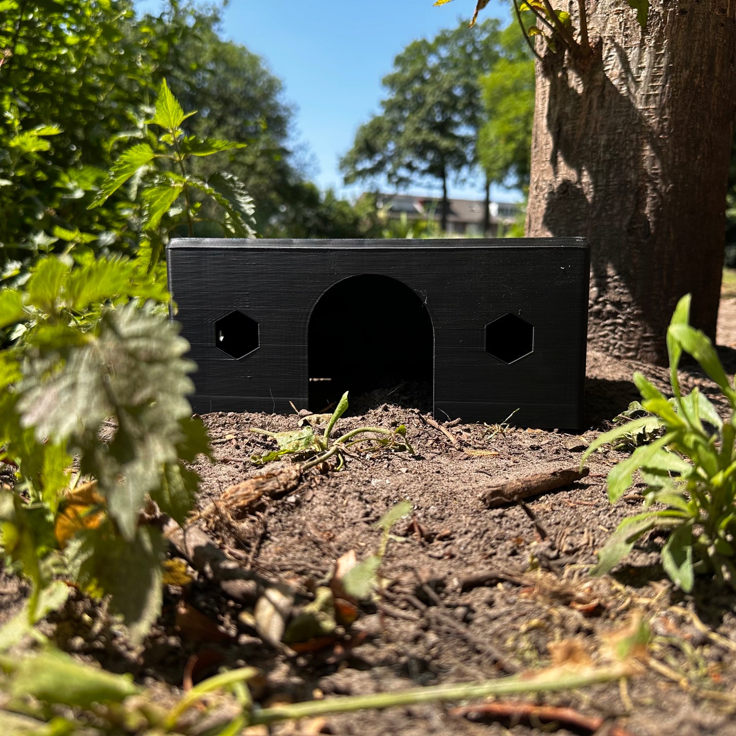 HexaHouse van zwart kleurig PLA, geplaatst in een natuurlijke buitenomgeving – 3D-geprint knaagdierhuisje met zeshoekige ramen.