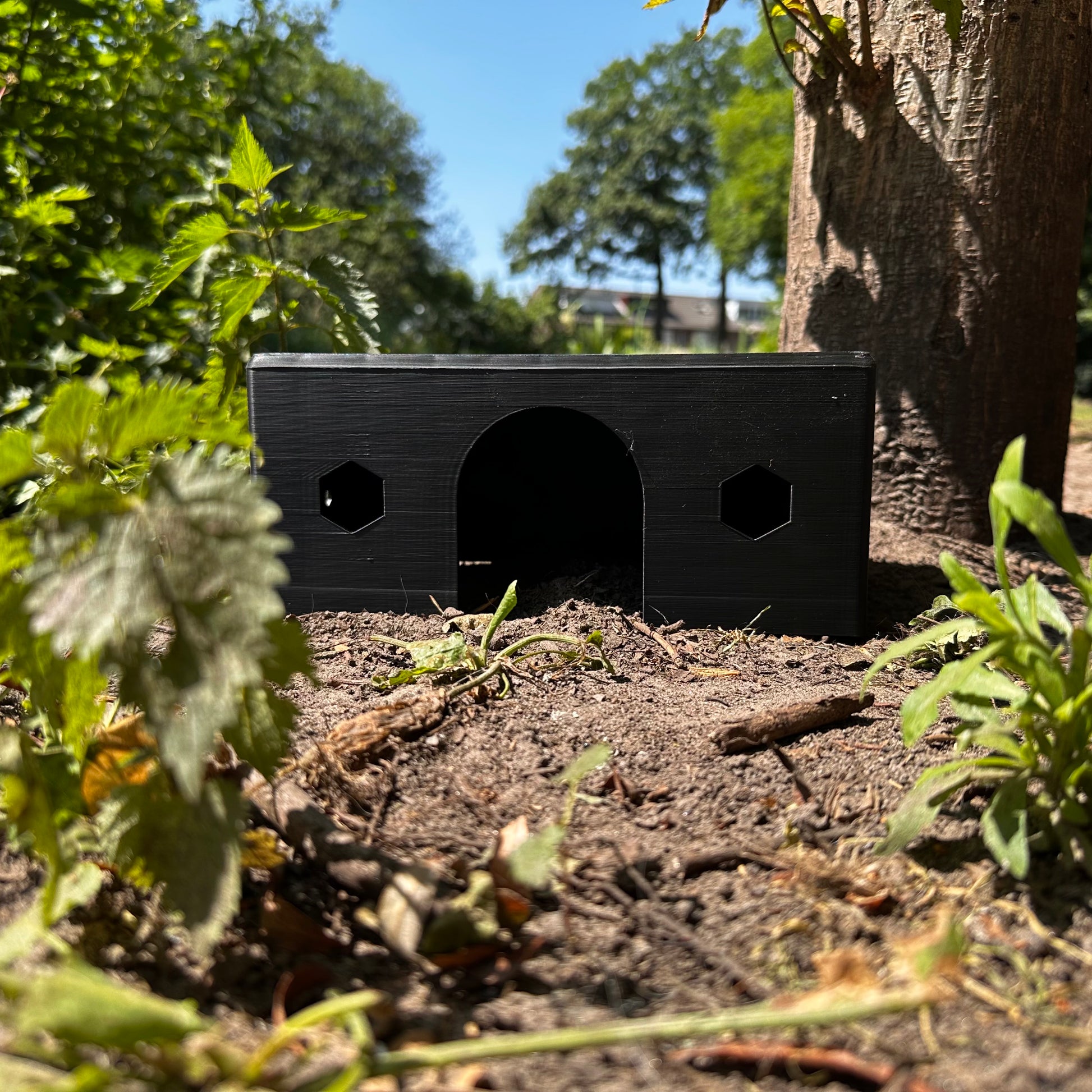 HexaHouse van zwart kleurig PLA, geplaatst in een natuurlijke buitenomgeving – 3D-geprint knaagdierhuisje met zeshoekige ramen.