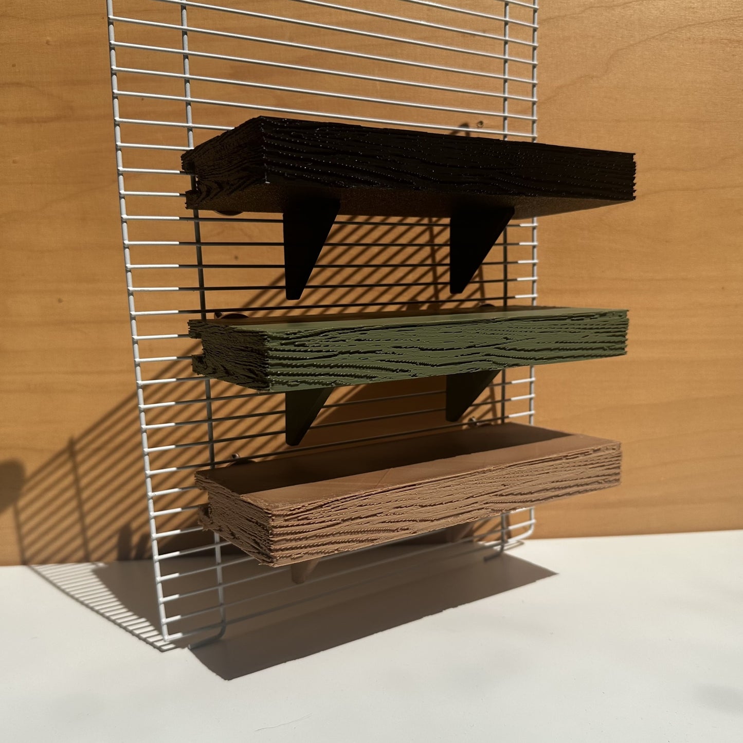 Verstelbare knaagdierplankjes in houtkleur, olijfgroen en zwart – als demonstratie bevestigd aan tralies op verschillende hoogtes.