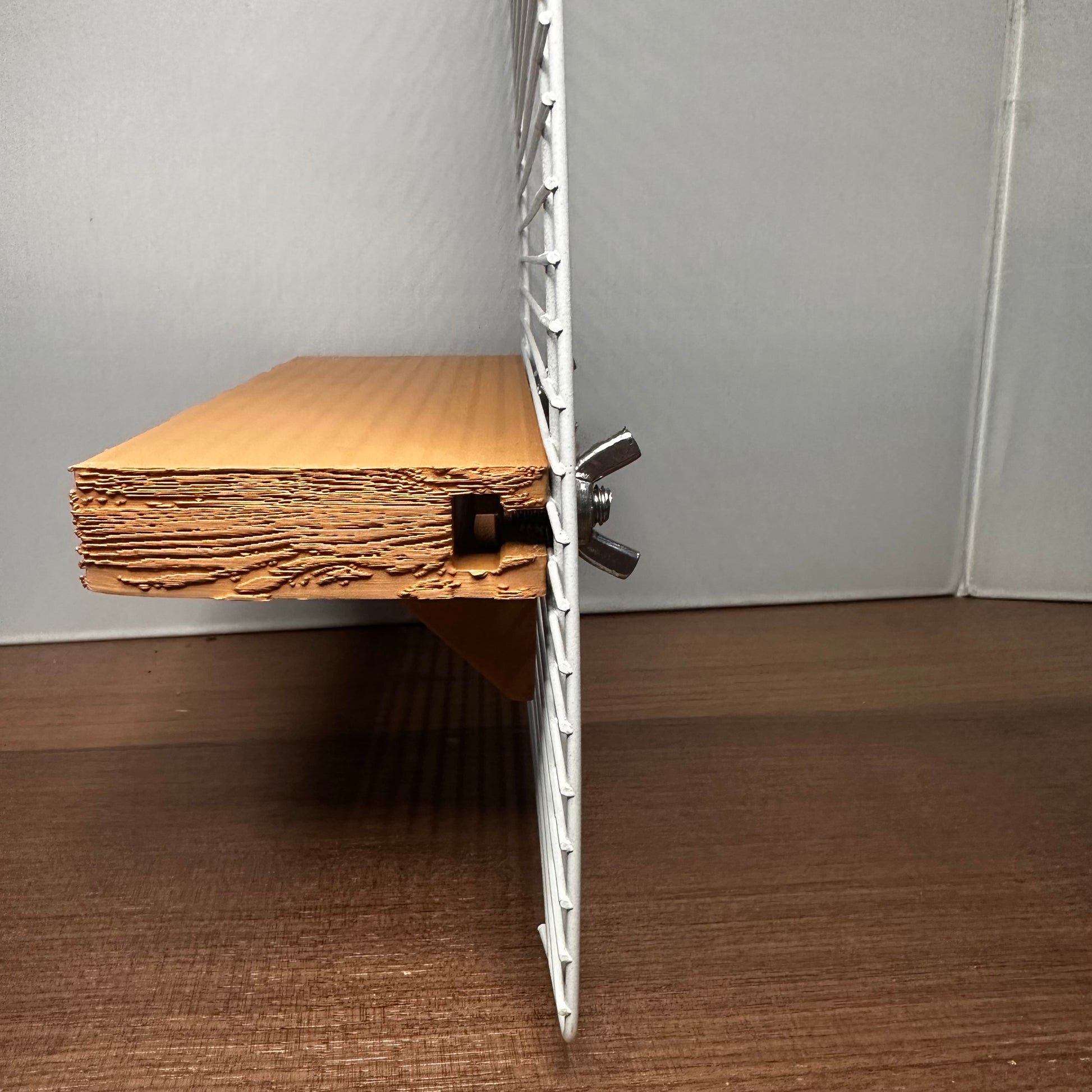 Zijaanzicht van een verstelbaar knaagdierplankje van houtkleurig PLA, bevestigd aan tralies – bevestigingssysteem en diepte goed zichtbaar.